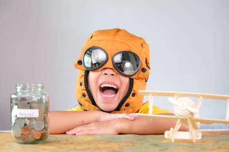 Smiling little girl in a pilot hat with coins in a clear jar for savings for travel and a wooden toy...
