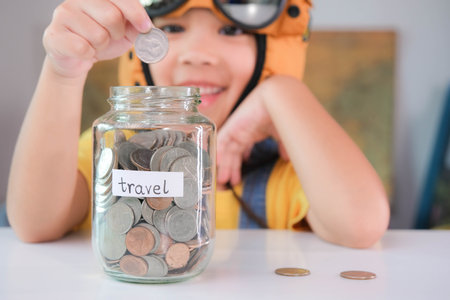 Cute little girl in a pilot hat is putting coins in a clear jar on the table to save for travel....
