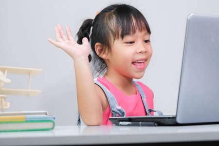 Smiling little Asian girl studying online having video call distant class with teacher using laptop. Happy little girl wave greeting with tutor while studying online with laptop at home. homeschoolingの写真素材
