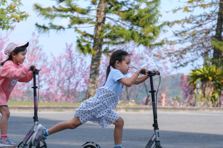 Active sisters riding scooters on street in outdoor park on summer day. Happy Asian kids riding kick...
