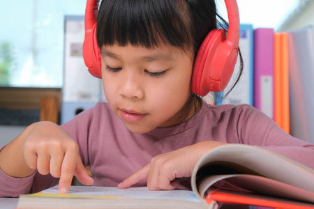 Cheerful little girl in headphones reading a book sits at the table in her room at home. Technology and hobbies for childrenの写真素材
