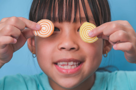 Smiling cute little girl covering her eyes with two gelatinous sweets isolated on blue background.  Children eat sugary sweets, causing loss teeth or tooth decay and unhealthy oral care.の写真素材