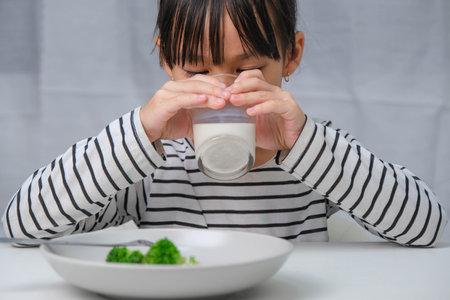 Cute Asian girl drinking a glass of milk in the morning before going to school. Little girl eats healthy vegetables and milk for her meals. Healthy food in childhood.の写真素材