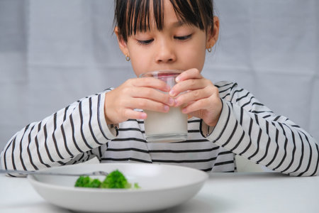 Cute Asian girl drinking a glass of milk in the morning before going to school. Little girl eats healthy vegetables and milk for her meals. Healthy food in childhood.の写真素材
