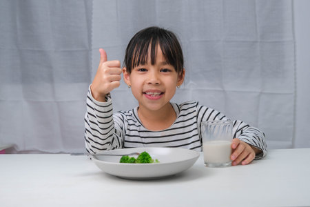 Cute Asian girl drinking a glass of milk in the morning before going to school. Little girl eats healthy vegetables and milk for her meals. Healthy food in childhood.の写真素材