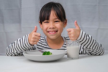 Cute Asian girl drinking a glass of milk in the morning before going to school. Little girl eats healthy vegetables and milk for her meals. Healthy food in childhood.の写真素材