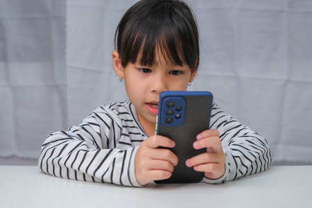 Cute little girl using smartphone and smiling while sitting at table in house. Happy Asian girl holding phone in hands, online learning or homeschooling, listening to music or playing games.の写真素材