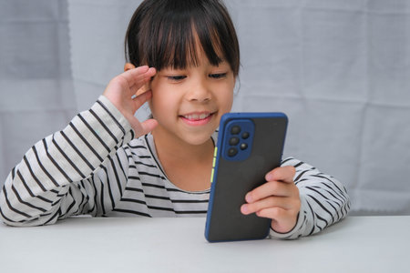 Cute little girl using smartphone and smiling while sitting at table in house. Happy Asian girl holding phone in hands, online learning or homeschooling, listening to music or playing games.の写真素材