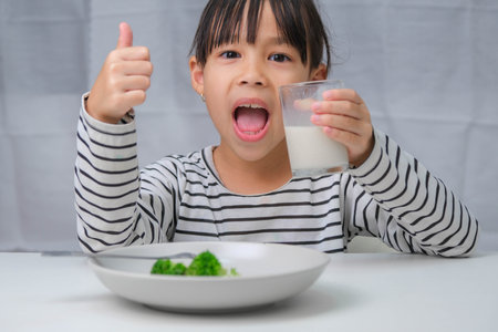 Cute Asian girl drinking a glass of milk in the morning before going to school. Little girl eats healthy vegetables and milk for her meals. Healthy food in childhood.の写真素材