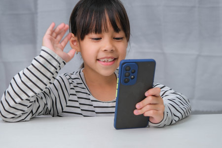 Cute little girl using smartphone and smiling while sitting at table in house. Happy Asian girl holding phone in hands, online learning or homeschooling, listening to music or playing games.の写真素材