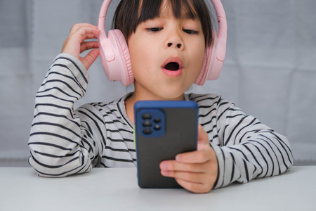 Cute elementary school girl wearing headphones holding a smartphone. Happy Asian girl studying online on smartphone or homeschooling, listening to music or playing games.の写真素材