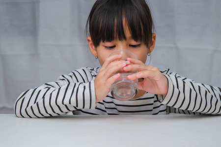 Cute little girl drinking water from glass on white curtain background. Healthy lifestyle and refreshment concept.の写真素材