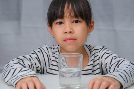 Cute little girl drinking water from glass on white curtain background. Healthy lifestyle and refreshment concept.の写真素材