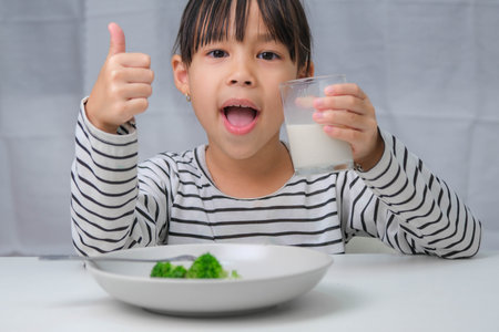 Cute Asian girl drinking a glass of milk in the morning before going to school. Little girl eats healthy vegetables and milk for her meals. Healthy food in childhood.の写真素材