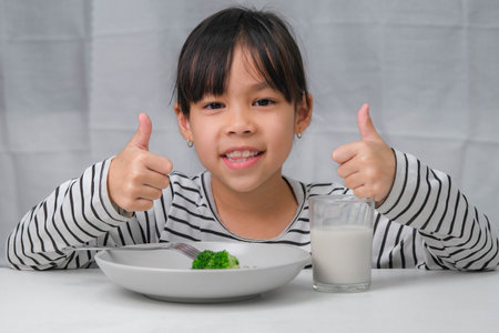 Cute Asian girl drinking a glass of milk in the morning before going to school. Little girl eats healthy vegetables and milk for her meals. Healthy food in childhood.の写真素材