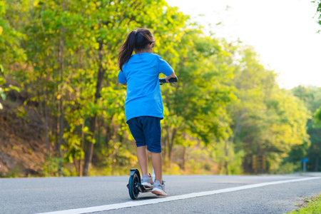Active little girl riding a scooter on the road in the park on a summer day. Cute little girl wearing helmet enjoys riding kick scooter in outdoor park. Leisure activities and outdoor sports for children.の写真素材