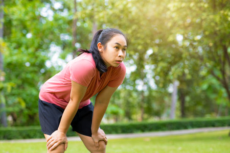 Young Asian woman standing bent over and breathing after running in the park. Young healthy woman taking a break after running. Healthy lifestyle concept.の写真素材