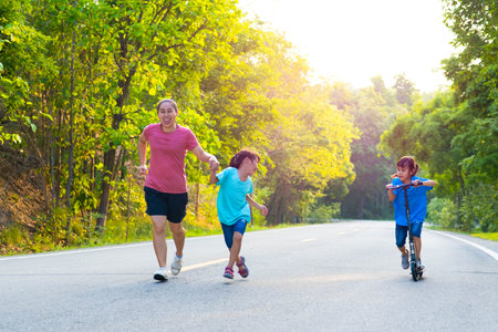 Active little girl riding a scooter on the street in the park on a summer day with her mother and sister jogging. Family spending time together.の写真素材