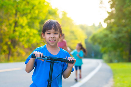 Active little girl riding a scooter on a park road on a summer day against her mother and sister in a blurred background. Family spending time together.の写真素材