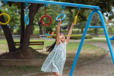 Happy girl hanging on monkey bar by hand doing exercise. Little Asian girl playing at outdoor playground in the park on summer vacation. Healthy activity.の写真素材