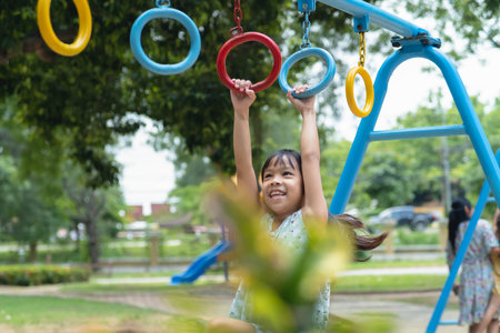 Happy girl hanging on monkey bar by hand doing exercise. Little Asian girl playing at outdoor playground in the park on summer vacation. Healthy activity.の写真素材