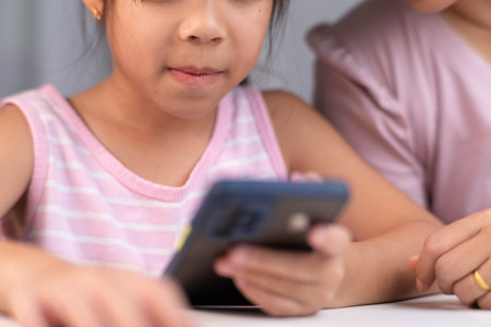 Happy Asian family mother and cute daughter using phone together at home. Mother and little girl laughing happily using smartphone watching funny social media video app sitting at table together.の写真素材
