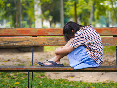 A sad little girl is sitting alone on bench in park. The little girl is unhappy and anxious because she has no one to play with.の写真素材
