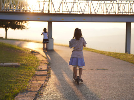 Cute sisters riding scooters in a park during a summer sunset in the park. Active leisure and outdoor sport for child.の写真素材