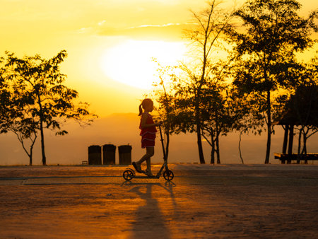 Silhouette of a cute little girl riding a scooter in a park during a summer sunset at a public park. Active leisure and outdoor sport for child.の写真素材