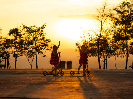 Silhouette of cute sisters riding scooters in the park during a summer sunset at a public park. Active leisure and outdoor sport for child.の写真素材