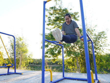 Asian adult woman exercising with outdoor fitness equipment in a park.の写真素材