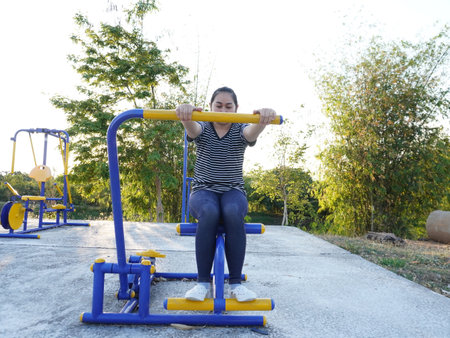 Asian adult woman exercising with outdoor fitness equipment in a park.