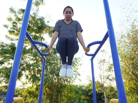 Asian adult woman exercising with outdoor fitness equipment in a park.の写真素材