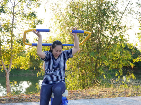 Asian adult woman exercising with outdoor fitness equipment in a park.の写真素材