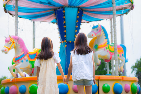 Rear view of two Asian girls having fun walking in an amusement park, holding hands and laughing while enjoying family vacation.の写真素材