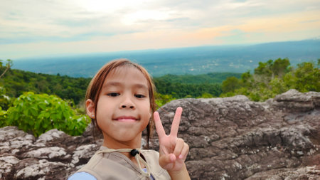 Cute girl smiles and makes a victory gesture looking at the camera while walking on a mountain, family trip in mountains. Mother and children are enjoying nature outdoors.の写真素材
