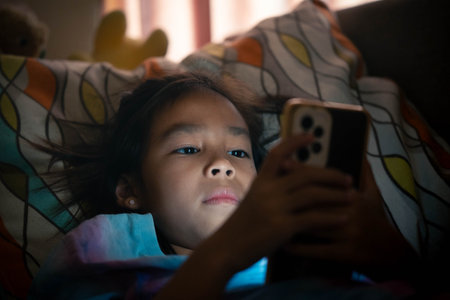 An Asian girl is looking at her mobile phone in low light and the blue light from the phone is...