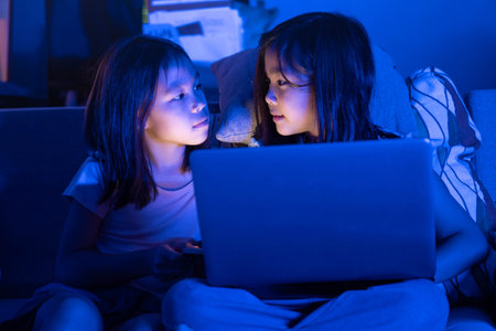 Two Asian girls sit on the sofa watching a laptop at night. Parental control, screen addiction, and bedtime behavior.の写真素材