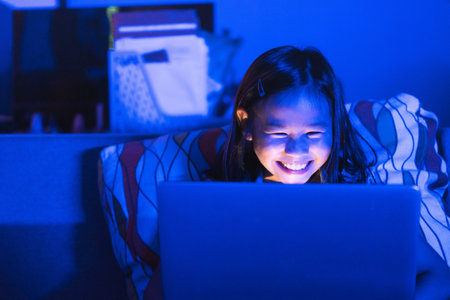 Asian girl sitting on the couch watching laptop at night. Parental control, screen addiction, and bedtime behavior.の写真素材