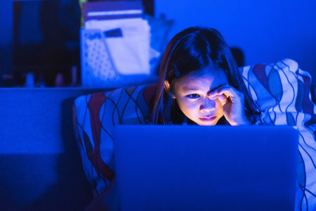 Asian girl sitting on the couch watching laptop at night. Parental control, screen addiction, and bedtime behavior.の写真素材