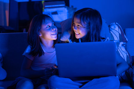 Two Asian girls sit on the sofa watching a laptop at night. Parental control, screen addiction, and bedtime behavior.の写真素材