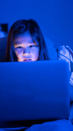 Asian girl sitting on the couch watching laptop at night. Screen addiction and bedtime behavior.
