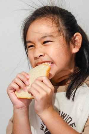 An Asian girl smiling while biting into a slice of bread, showing a playful and happy expression...