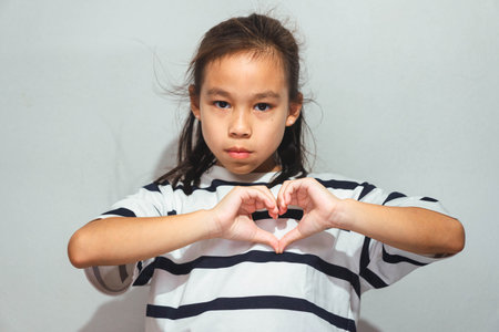 An Asian girl making a heart shape with her hands, symbolizing love, care, and friendship, standing...