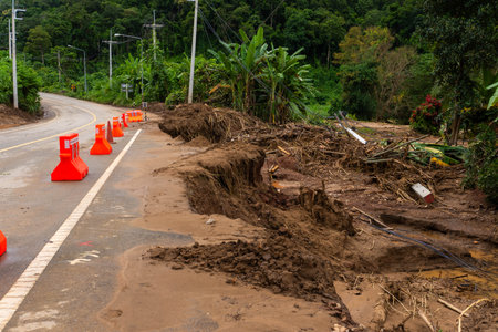 Rural road severely damaged by flood and landslide, with mud, soil erosion, and orange barriers placed for safety and warning.の写真素材