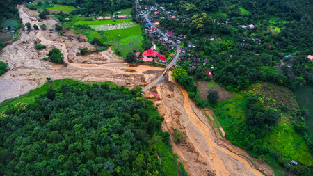 Drone aerial view of muddy floodwaters cutting through farmland and reaching a rural village, showing the dramatic contrast between lush green fields and disaster destruction.の写真素材