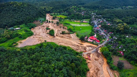 Drone aerial view of muddy floodwaters cutting through farmland and reaching a rural village, showing the dramatic contrast between lush green fields and disaster destruction.の写真素材