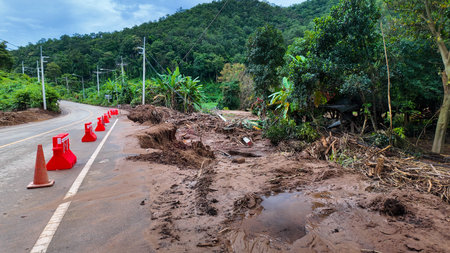 A damaged mountain road eroded by flood and landslide, with warning cones and barriers placed for safety after heavy rain.の写真素材
