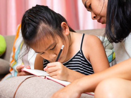An Asian mother helping her daughter to write in a notebook at home. The image shows family bonding, learning, and education in daily life, representing parental support, childhood development, and love in the family.の写真素材