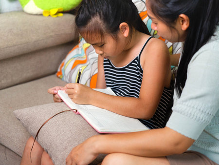 An Asian mother helping her daughter to write in a notebook at home. The image shows family bonding, learning, and education in daily life, representing parental support, childhood development, and love in the family.の写真素材